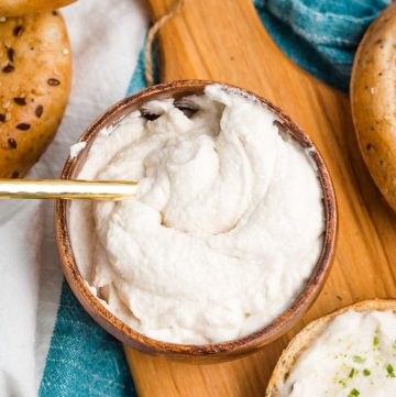 A bowl of vegan cream cheese on a wooden cheese board, surrounded by bagels.